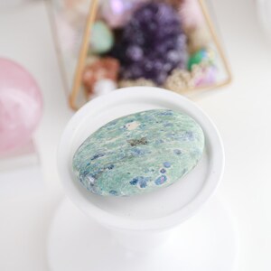 May include: A smooth, oval-shaped green and blue gemstone with a speckled pattern. The stone is resting on a white pedestal.