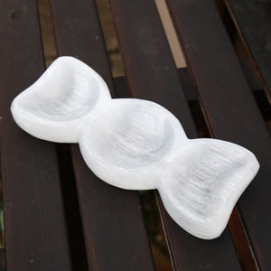 May include: A white selenite crystal bowl with three rounded sections. The bowl is carved from a single piece of selenite and has a smooth, polished finish.