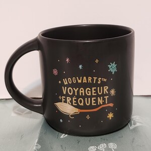 May include: Black ceramic mug with a gold Hogwarts Frequent Flyer design. The mug features a broomstick and stars.
