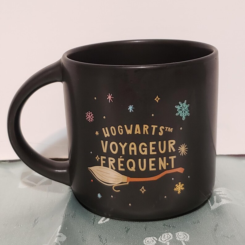 May include: Black ceramic mug with a gold Hogwarts Frequent Flyer design. The mug features a broomstick and stars.