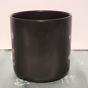 May include: A black ceramic mug with a colorful design. The mug has a wide rim and a rounded bottom.