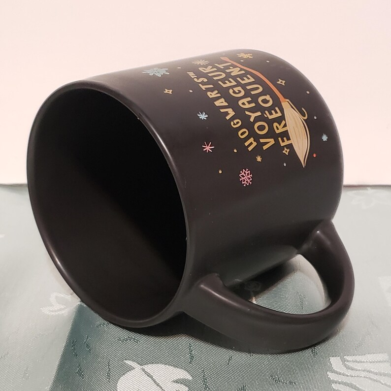 May include: Black ceramic mug with a gold design that says "Hogwarts&trade; Voyageur Frequent" with a broom and snowflakes.
