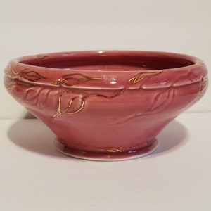 May include: A pink ceramic bowl with a gold leaf design. The bowl has a wide rim and a flared base.