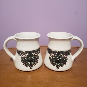 May include: Two white ceramic mugs with a brown and green floral design. The mugs have a rounded shape and a handle.