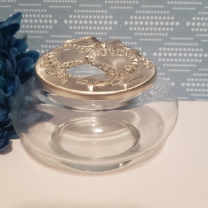 May include: A clear glass jar with a silver lid featuring two intertwined hearts with floral accents.