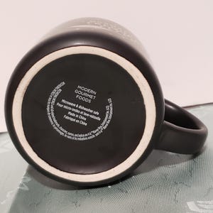 May include: Black ceramic mug with a white Modern Gourmet Foods logo and text that reads "Microwave & dishwasher safe. Pour micro-ondes et lave-vaisselle. Made in China. Fabriqu&eacute; en Chine."
