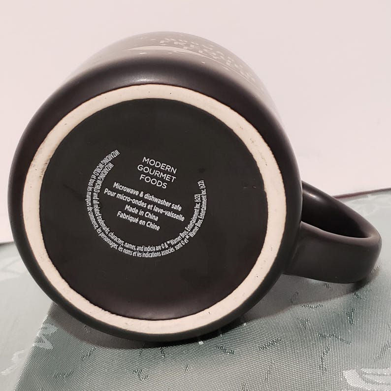 May include: Black ceramic mug with a white Modern Gourmet Foods logo and text that reads "Microwave & dishwasher safe. Pour micro-ondes et lave-vaisselle. Made in China. Fabriqu&eacute; en Chine."