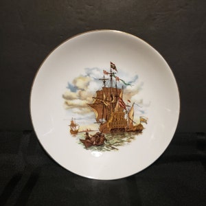 May include: A white ceramic plate with a gold rim features a vintage illustration of a large sailing ship with billowing sails and multiple flags. The ship is surrounded by smaller boats and a cloudy sky.