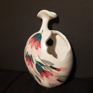 May include: White ceramic wall vase featuring a floral design. The vase has a distinctive shape with a cut-out opening and a small spout. The flowers are painted in shades of pink, red, and green.