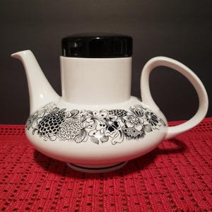 May include: White teapot with a black floral design and a black lid. The teapot has a round body and a spout on the side.