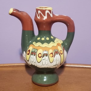 Peut inclure: Un petit pichet en céramique vert, brun et blanc avec un design unique. Le pichet a un bec verseur et une anse. Il est décoré de volutes et de points jaunes, verts et blancs.