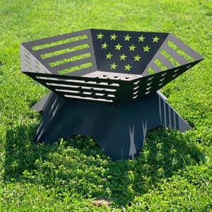 Puede incluir: Un pozo de fuego de metal negro con forma de estrella con un diseño recortado de la bandera estadounidense. El pozo de fuego está sentado en un césped.