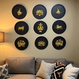 May include: Nine black circular wall decorations with gold relief designs. The designs include an elephant, a lotus flower, a bull, a vase, and a mythical bird. The decorations are mounted on a beige wall above a gray sofa.