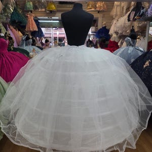 May include: A white crinoline petticoat with multiple hoops, displayed on a black mannequin. The petticoat is made of layers of tulle and has a lace trim at the bottom. Other dresses are visible in the background.