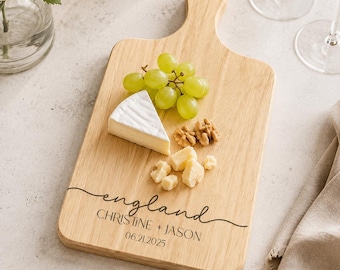 Könnte beinhalten: Ein helles Holz-Servierbrett mit Griff und Aufhängeloch. Das Brett ist mit dem eingravierten Text "england", "CHRISTINE + JASON" und "06.21.2025" versehen. Es ist mit einem Stück Brie-Käse, grünen Weintrauben und Walnüssen belegt.