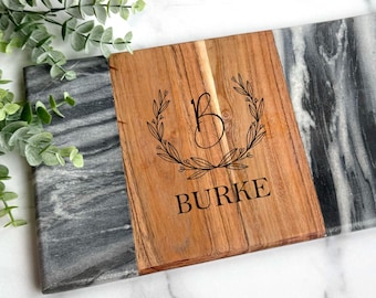 Könnte beinhalten: Ein rechteckiges Servierbrett mit Holz- und Marmordesign. Der Mittelteil ist aus Holz, graviert mit dem Buchstaben "B" in einem Kranz und dem Namen "BURKE". Die Marmorteile sind grau und weiß. Grünzeug befindet sich in der oberen linken Ecke.