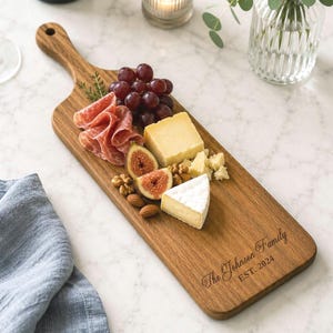 May include: A rectangular wooden serving board with a handle, featuring the engraved text The Johnson Family EST. 2024. The board is arranged with a selection of red grapes, sliced figs, wedges of cheese, cured meat, and walnuts.