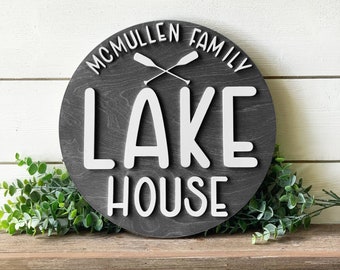 Könnte beinhalten: Ein rundes Holzschild mit dem Text "MCMULLEN FAMILY LAKE HOUSE" in weißen Buchstaben. Das Schild hat einen dunkelgrauen Hintergrund und zwei gekreuzte Ruder im Zentrum.