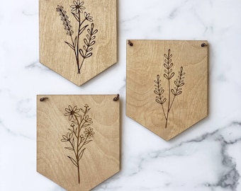 Könnte beinhalten: Drei Holz-Wanddekorationen mit floralem Design. Jedes Stück ist in Form eines Wimpels geformt und hat eine braune Schnur zum Aufhängen.