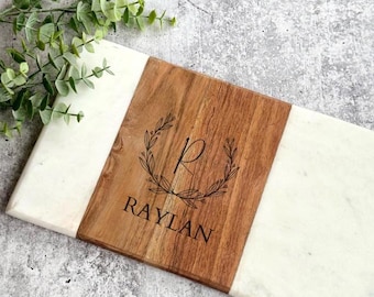 Könnte beinhalten: Ein rechteckiges Servierbrett mit weißer Marmorbasis und einem Mittelteil aus Holz. Der Holzabschnitt ist mit dem Namen "RAYLAN" und einem dekorativen Kranz graviert. Ein Zweig grüner Blätter befindet sich im Hintergrund.