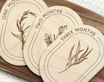 Könnte beinhalten: Drei Meilensteine für Babys aus Holz mit eingravierten botanischen Designs. Jeder ovale Marker ist mit einem Monat beschriftet: "ONE MONTH", "TWO MONTHS" und "THREE MONTHS". Die Marker sind aus hellem Holz und liegen auf einer Holzoberfläche.