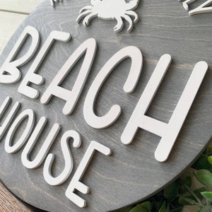 May include: A round wooden sign with a gray background and white lettering. The sign says "McMullen Family Beach House" with a white crab silhouette in the upper center of the sign.