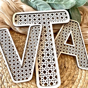 May include: Wooden letters spelling out the word "VATA" with a woven cane design. The letters are light-colored and sit on a woven basket. Green leaves and a tan cloth are in the background.