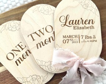 Könnte beinhalten: Drei Meilenstein-Holztafeln mit floralen Akzenten und graviertem Text. Eine Tafel lautet "ONE mon", eine andere "TWO mon", und die dritte zeigt "Lauren Elizabeth" mit Geburtsdaten: 7. März 2023, 3,18 kg, 48,26 cm, 16:46 Uhr. Ein rosa Band ist an den Tafeln befestigt.
