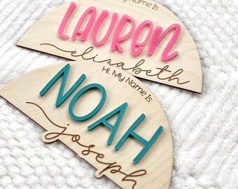 Könnte beinhalten: Zwei halbkreisförmige Namensschilder aus Holz. Eines hat den Namen "Lauren" in Rosa, darunter "Elizabeth". Das andere hat "Noah" in Türkis, darunter "Joseph". Beide haben "Hi. My Name Is" über den Namen.
