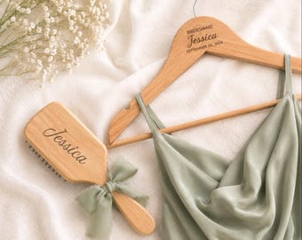 May include: A light wood clothes hanger engraved with the text BRIDESMAID Jessica SEPTEMBER 06, 2024, a light wood hairbrush engraved with the name Jessica and tied with a sage green ribbon, and a sage green satin dress with thin straps.