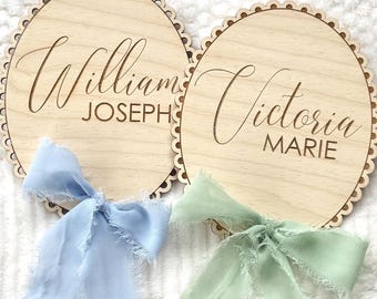 Könnte beinhalten: Zwei runde Namensschilder aus Holz mit gewellten Kanten. Auf einem Schild steht "William JOSEPH" und auf dem anderen "Victoria MARIE". Jedes Schild hat eine Stoffschleife, eine hellblaue und eine hellgrüne.