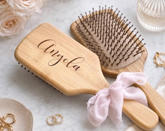 May include: A rectangular bamboo hairbrush with the name Angela engraved in a cursive script on the handle. The brush features dark brown bristles and is tied with a light pink fabric ribbon. The natural wood grain is visible on the surface.