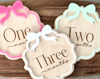 Könnte beinhalten: Meilenstein-Tafeln aus Holz für die ersten drei Monate des Babys. Jede Tafel hat eine dekorative Schleife und die Monatszahl in einer stilvollen Schriftart. Die Ränder sind in Rosa, Weiß und Mintgrün bemalt.