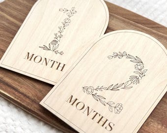 Könnte beinhalten: Zwei Meilenstein-Tafeln aus Holz mit gewölbten Oberteilen. Die Tafeln sind helles Holz mit eingravierten Blumendesigns und den Wörtern "1 MONAT" und "2 MONATE". Die Tafeln liegen auf einer Holzoberfläche.