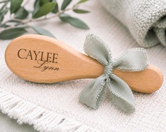 May include: A light-colored wooden hairbrush with a smooth, oval-shaped handle. The name CAYLEE Lynn is engraved in dark lettering on the handle. A sage green fabric ribbon with frayed edges is tied into a bow around the neck of the brush handle.