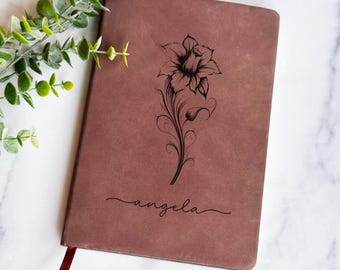 Könnte beinhalten: Ein braunes Notizbuch mit einem Blumendesign und dem Namen "angela" in Schreibschrift. Das Notizbuch hat ein rotes Band als Lesezeichen und liegt auf einer weißen Marmoroberfläche mit grünem Laub.