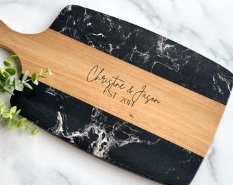 May include: A rectangular serving board featuring a central light brown wood panel flanked by two black marble-patterned sections. The wood center is engraved with the text "Christine & Jason" and "EST. 2014" in a black script and sans-serif font.