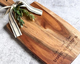Könnte beinhalten: Ein Holzbrett zum Schneiden mit einem Griff, graviert mit dem Text "Home Sweet Home" und den Koordinaten "32.00035° N, 80.9737° W Wilmington, North Carolina". Das Brett ist mit einem weiß-schwarz gestreiften Band und einem Zweig Grünzeug gebunden.