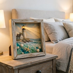 May include: A square shadow box frame with a distressed wood finish containing a coastal scene. The artwork features a lighthouse with a glowing light, a small house on a rocky cliff, and a foreground filled with blue sea glass and seashells.