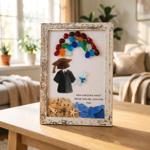 Può includere: Un'opera d'arte incorniciata con vetro marino che forma un arcobaleno, un tocco e una toga da laurea. Sono inclusi una piccola pergamena in vetro marino, una conchiglia e una stella marina. Il testo recita: NEW HORIZONS AWAIT DREAM. EXPLORE DISCOVER. 2026.