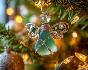 May include: A handcrafted Christmas ornament in the shape of an angel. The angel features a green glass body, wire wings, and a clear glass head. It is suspended from a Christmas tree branch, with warm lights in the background.