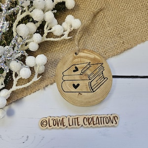 May include: A wooden ornament shaped like a stack of books with hearts on the covers. The ornament is decorated with a black outline and is hanging from a string. The text "OLOWE LITE CREATIONS" is printed on a separate wooden piece.