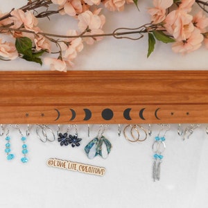 May include: Wooden earring holder with a moon and star design. The holder has hooks for hanging earrings. There are several pairs of earrings hanging on the holder, including blue crystal earrings, silver hoop earrings, and black flower earrings. The holder has the text "@LOWE LITE CREATIONS" on the bottom.