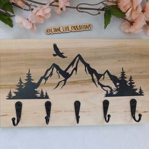 Peut inclure: Porte-clés en bois avec un motif de montagne et d'aigle en silhouette. Il possède quatre crochets en métal noir. Le texte "OLOWE LITE CREATIONS" est visible en haut du porte-clés.