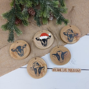 May include: Four wooden ornaments featuring painted cow faces. One cow is wearing a red Santa hat. The ornaments are displayed on a hessian background. The text "OLOWE LITE CREATIONS" is visible in the bottom right corner.