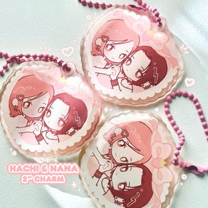 May include: Three pink acrylic charms featuring a cartoon illustration of two girls embracing. Each charm is 2 inches in diameter and has a pink beaded chain. The text "Hachi & Nana 2" Charm" is visible on the image.