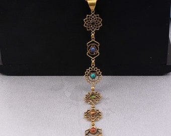 May include: A gold-colored necklace featuring seven decorative pendants, each with a different colored gemstone. The pendants are arranged vertically and connected by a delicate chain. The necklace is displayed against a black background.