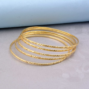 May include: A set of six gold-colored bangles with a textured, etched design. The bangles are thin and circular, and they are arranged in a slightly overlapping pattern. The background is a light gray surface with a blue backdrop.