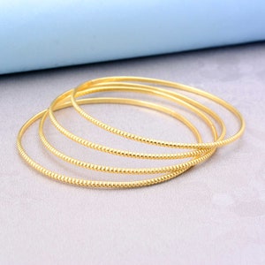 Puede incluir: Cuatro pulseras doradas, cada una con un acabado liso y pulido. Una pulsera tiene un patrón texturizado y punteado. Las pulseras están dispuestas de forma ligeramente superpuesta, creando un efecto de capas. El fondo es de color neutro.