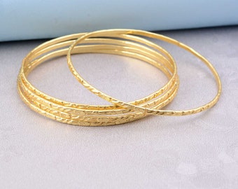 Könnte beinhalten: Eine Sammlung goldfarbener Armreifen. Die Armreifen haben eine strukturierte Oberfläche und variieren in der Dicke. Der Schmuck wird auf einer hellgrauen Oberfläche mit hellblauem Hintergrund präsentiert.